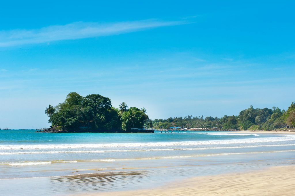 A serene view of the beach in Mirissa, Sri Lanka with gentle waves and a lush island under a bright blue sky.