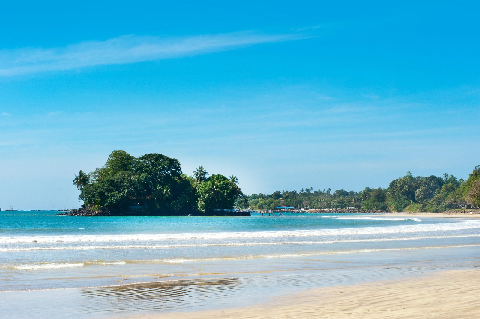 A serene view of the beach in Mirissa, Sri Lanka with gentle waves and a lush island under a bright blue sky.